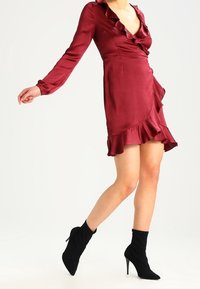 Robe enveloppante en satin bourgogne avec des manches longues, un ourlet volanté et un décolleté en V profond ; assortie à des bottines noires à talons aiguilles.