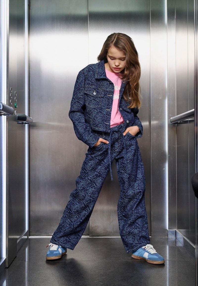 Mornarsko-modra denim obleka z vzorcem cvetja, obrezana jakna, ohlapne hlače z zavezanim pasom in modri supergi z belimi poudarki.