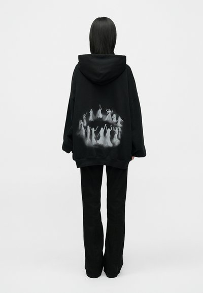 Fekete, túlméretezett kapucnis pulóver, amelynek hátán fehér táncoló alakok grafikai terve látható, kenguruzsebbekkel és húzózsinóros kapucnival.