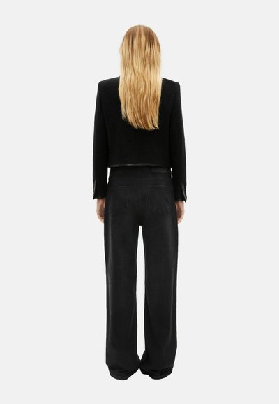 Giacca nera corta con tessuto strutturato, abbinata a pantaloni neri a taglio ampio. Capelli lunghi e ondulati visibili sul retro.