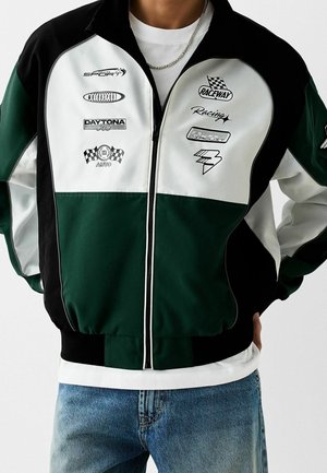 Osoba oblečená v zelené, černé a bílé závodní bundě s motorsportovými logy přes bílé tričko a modré džíny.