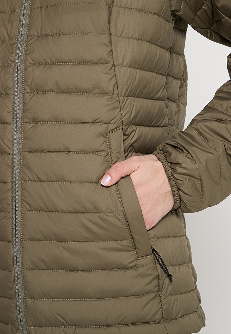 Ręka spoczywająca wewnątrz bocznej kieszeni brązowej, pikowanej kurtki zapinanej na zamek z elastycznymi mankietami.