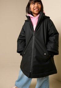 Juodas neperšlampamas paltas su užtrauktuku priekyje, plačiomis rankovėmis, šoninėmis kišenėmis ir gaubtu; dėvimas virš rausvo džemperio su kapišonu ir šviesiai mėlynų džinsų.