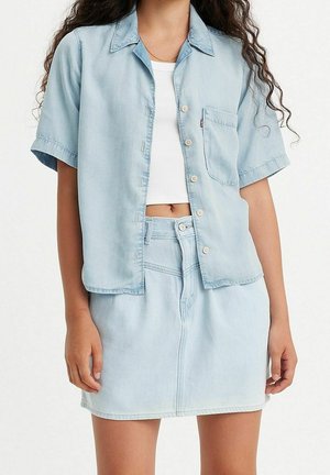Femme portant une chemise boutonnée à manches courtes bleu clair sur un crop top blanc, associée à une jupe en jean bleu clair assortie.