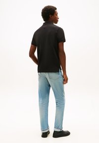 Černé polo tričko s krátkými rukávy, zobrazené na osobě nosící světle modré džíny a černé boty, zobrazený zezadu.