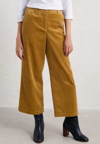 Pantalons larges en velours côtelé jaune moutarde, avec des côtes verticales, une fermeture à bouton et des poches avant, associés à des bottines noires.