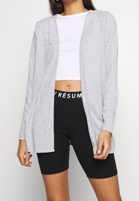 Cardigan aperto grigio chiaro realizzato in morbido tessuto, con maniche lunghe e vestibilità comoda. Abbinato a un top corto bianco e leggings neri.