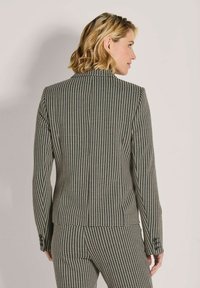 Frau mit kurzem blonden Haar, die einen schwarz-weißen karrierten Blazer und passende Hose trägt, seitlich vor einem schlichten Hintergrund stehend.