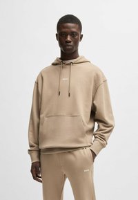 Beige hoodie with a front pocket and drawstring hood; features white brand logo "BOSS" on the chest. Matched with beige track pants.
