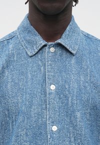 Világoskék farmering texturált felülettel. Hegyes gallérral és fehér gombokkal rendelkezik. Közeli kép, amely az elejére fókuszál.