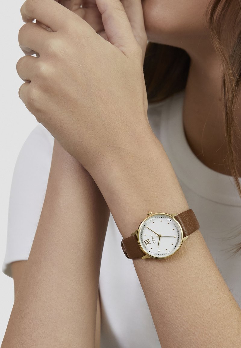 Montre à bracelet en cuir marron avec un cadran blanc, des accents dorés, des marqueurs numériques et un bracelet texturé. Design simple et élégant.