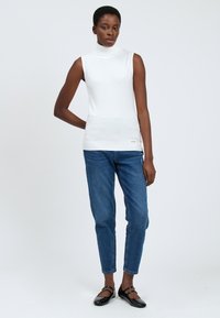Top bianco senza maniche a collo alto abbinato a jeans blu. Il top presenta un orlo a coste, mentre le scarpe sono ballerine nere con una finitura lucida.