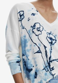 Weißer V-Ausschnitt-Pullover mit blauem Blumenmuster und spritzenden Farben. Bietet einen lockeren Schnitt, lange Ärmel und gerippte Bündchen mit blauen Akzenten.