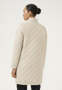 Hellbeige gesteppter Mantel mit hohem Kragen, langen Ärmeln und diagonaler Nahtführung, der bis zur Mitte des Oberschenkels reicht. Minimalistisches Design.