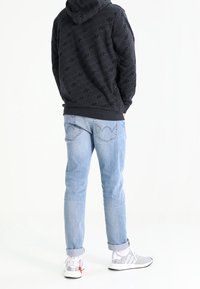 Osoba v černé vzorované mikině s kapucí, světle modrých ohrnutých jeansách a bílých teniskách, stojí zády k bílé zdi.