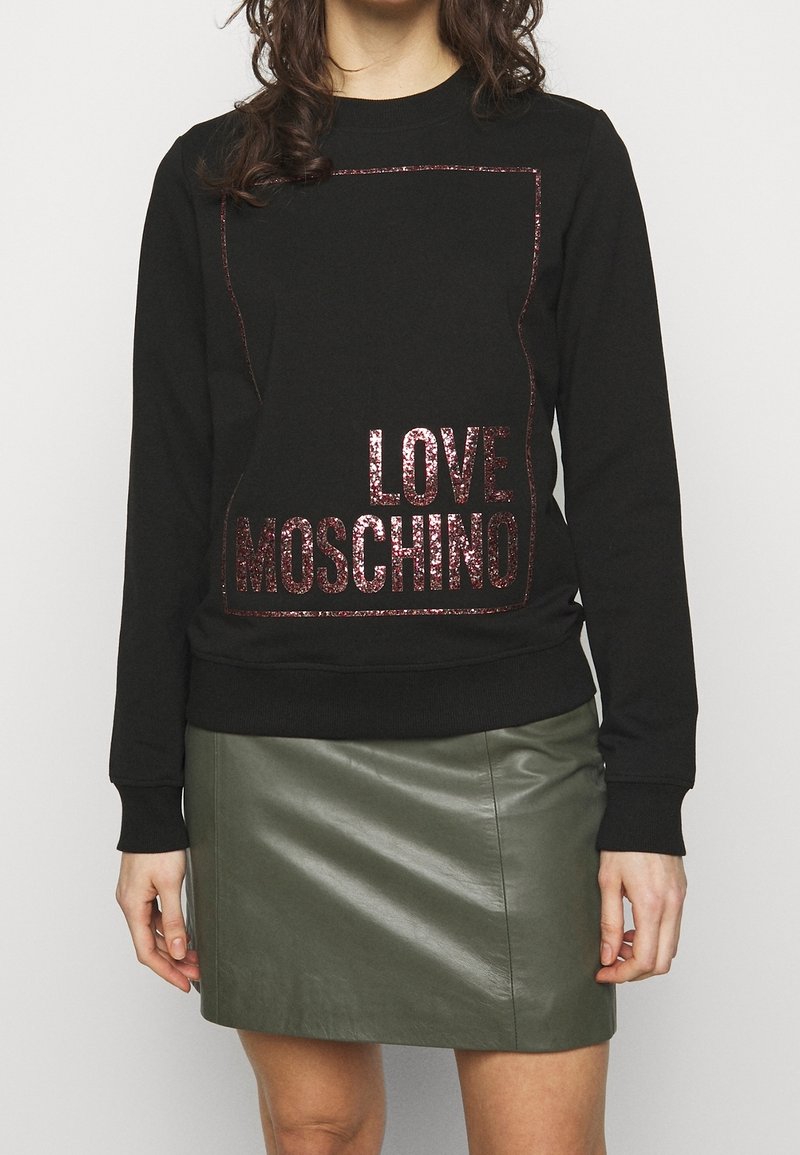 Černá mikina se třpytivým nápisem "LOVE MOSCHINO", čtvercovým okrajem a žebrovanými lemy, nošena přes zelenou koženou mini sukni.