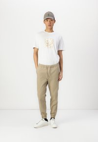 Camiseta blanca con un patrón cuadrado en color beige, joggers beige con puños elásticos y zapatillas blancas. Modelo lleva una gorra gris claro.