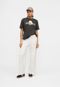 Γυναίκα που φοράει μαύρο μπλουζάκι με την επιγραφή «DREAMERS» και ένα λευκό λουλούδι, λευκό παντελόνι με φαρδύ πόδι, μαύρα και λευκά αθλητικά παπούτσια και μπεζ καπέλο, μπροστά σε λευκό φόντο.