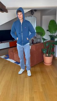 Blue zip-up hoodie, light gray pants, and white sneakers. Indoor setting with a brown couch and green plants in the background.