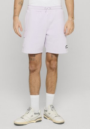 Man die lichtpaarse korte broek met zijzak, witte sokken en wit-grijze sneakers draagt, staand tegen een effen achtergrond.