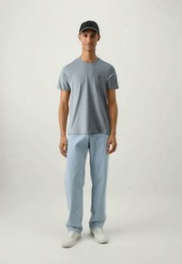 Svetlomodré bavlnené tričko s zeleným logom, spárované so svetlomodrými džínami. Doplnky zahŕňajú čiernu čiapku a biele tenisky.