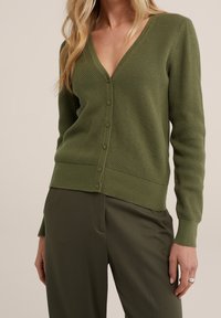 Olivově zelený svetr s výstřihem do V, texturovanou tkaninou a vroubkovanými manžetami a lemem. Disponuje zapínáním na knoflíky vpředu a dlouhými rukávy.