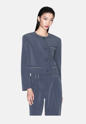 Frau, die einen grauen maßgeschneiderten Anzug mit großen Knöpfen und kontrastierenden weißen Nähten trägt, steht vor einem schlichten Hintergrund.