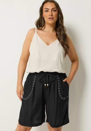 Vrouw met lang bruin haar, gekleed in een beige mouwloze top en zwarte short met trekkoord en witte stiksels, staand tegen een effen achtergrond.