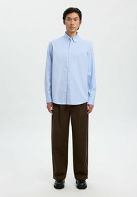 Man in een lichtblauw overhemd met knopen, donkere bruine wijde broek en zwarte schoenen, staand tegen een effen witte achtergrond.