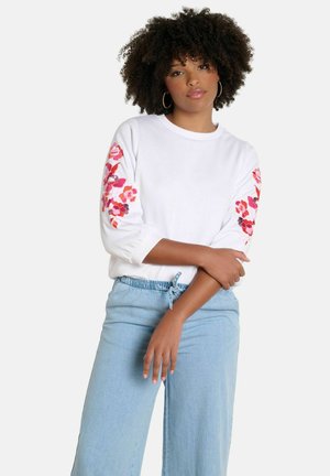 Woman with curly hair wearing white top with pink floral embroidery on sleeves and light blue drawstring pants, standing with arms crossed.