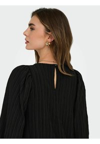 Black textured blouse featuring puffed sleeves and a keyhole back button detail. Lightweight fabric with vertical pleats. Gold hoop earrings.