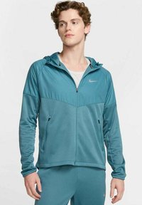 Chaqueta deportiva color azul verdoso con cremallera frontal, capucha y bolsillos laterales. Presenta una tela texturizada y un pequeño logo en el lado izquierdo del pecho.