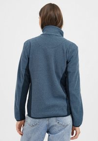 Veste en polaire texturé bleu avec un col haut, des panneaux latéraux sombres contrastants et un design de couture courbé à l'arrière.