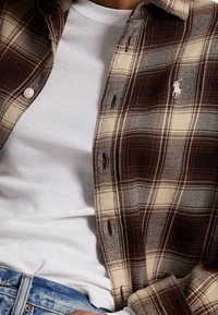 Brun och beige rutig flanellskjorta med knappar, bärs över en vit T-shirt. Blå denimjeans synliga vid midjan.