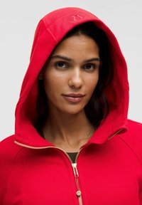 Červená mikina na zip s velkou kapucí, zlatým zipem a logem. Hladká látka s těsným střihem, zdůrazňující pohodlný atletický styl.