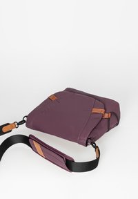Paarse stoffen tas met bruine leren accenten. Beschikt over een platte vorm, een verstelbare zwarte schouderband en een ritssluiting.