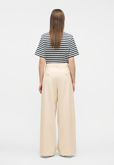 Γυναίκα με μακριά ίσια μαλλιά που φοράει μαύρο και λευκό ριγέ πουκάμισο και μπεζ παντελόνι με ψηλή μέση και φαρδιά πόδια, κοιτάζοντας προς τα πίσω.