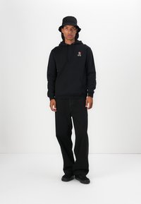 Felpa nera con ricamo di rose, vestibilità comoda, tasca a marsupio, abbinata a jeans neri a gamba larga e un cappello a secchiello nero. Sneakers nere completano il look.