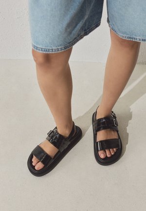 Person trägt einen knielangen Jeansrock und schwarze Lackledersandalen mit zwei Schnallen, steht auf hellem Boden.