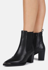 Botas de cuero negro hasta el tobillo con punta afilada, paneles elásticos laterales y tacón grueso. Presentan un acento metálico en la punta.