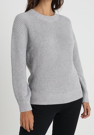 Mujer con un suéter de punto gris claro con puños y bajo acanalados, combinado con pantalones negros, posando contra un fondo liso.