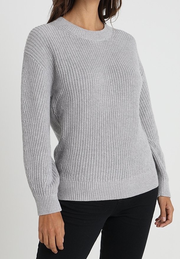 Femme portant un pull en maille gris clair avec poignets et ourlet côtelés, associé à un pantalon noir, posant devant un fond uni.