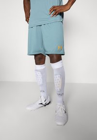 Light blue athletic shorts with orange Under Armour logo, white knee-high socks with grip, and white cleats with black accents.