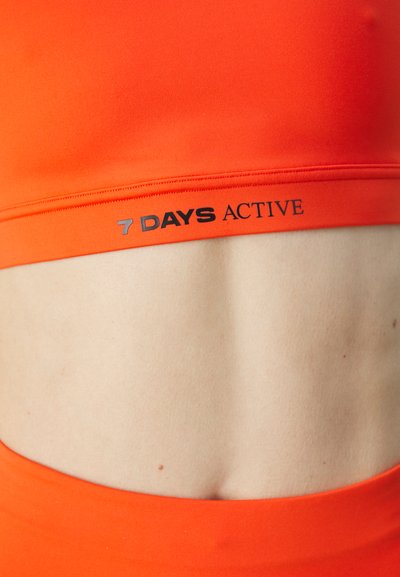 Bright orange activewear crop top with a smooth texture, featuring a black logo that reads "7 DAYS ACTIVE" along the bottom band.