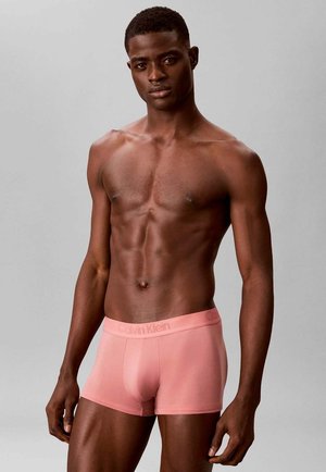 Shirtless man with muscular build wearing light pink Calvin Klein boxer briefs standing against a plain gray background.