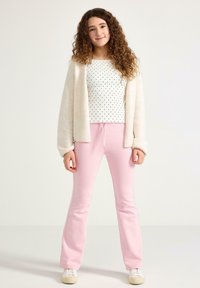 Witte top met polka dots, zachte beige cardigan en roze wijde broek met trekkoord. Model staat voor een neutrale achtergrond.