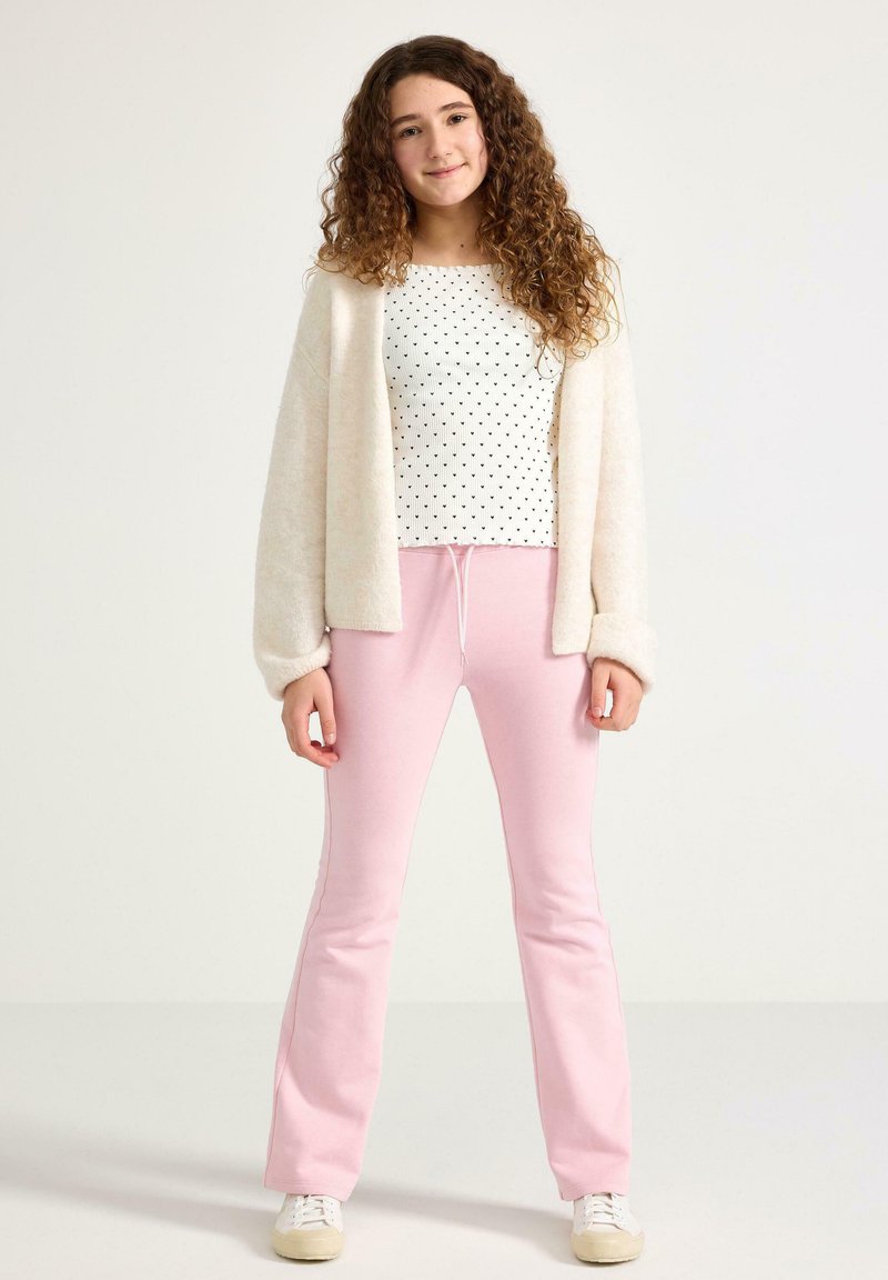 Witte top met polka dots, zachte beige cardigan en roze wijde broek met trekkoord. Model staat voor een neutrale achtergrond.