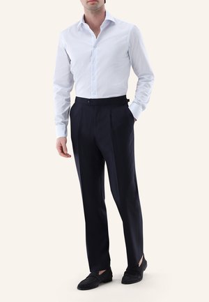 Mann trägt ein weiß gestreiftes Hemd, das in hoch taillierte schwarze Hose gesteckt ist, und schwarze Slipper, Hände entspannt.