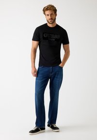 Czarna koszulka z krótkim rękawem z wytłoczonym logo "GUESS EST. 1981" z przodu, zestawiona z ciemnoniebieskimi dżinsami typu straight-leg i czarnymi trampkami z białą podeszwą.