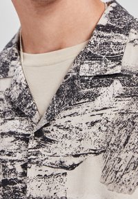 Zbliżenie na osobę w beżowej koszuli pod czarno-białą kurtką w abstrakcyjny wzór, z srebrnym naszyjnikiem.